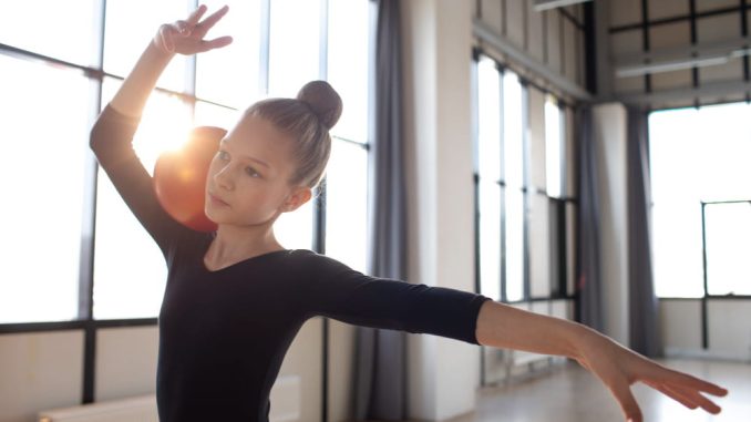 Une jeune gymnaste ou danseuse en justaucorps noir tient un ballon rouge contre son épaule tout en dansant, illustrant concrètement les bienfaits de la danse pour le développement de l'enfant et de l'ado.